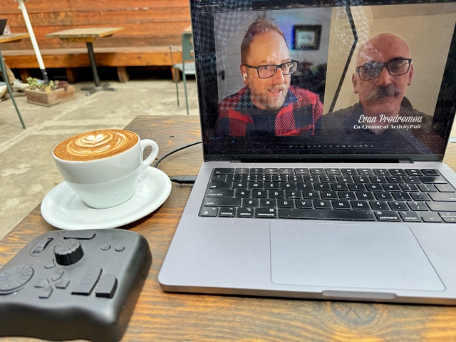 A laptop with video on screen of two men talking to each other. One is Doc, the other is Evan. 
There is a cappuccino and a controller on the table too. 