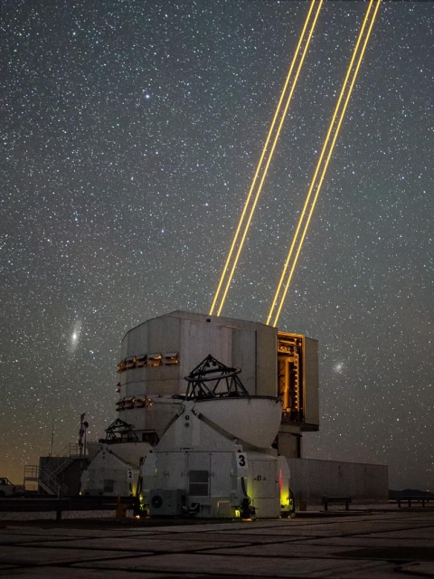 A long exposure image taken at night at an astronomical observatory, with several telescope domes. Four bright orange lines –– laser beams –– are coming out from one of them, against a starry backdrop.