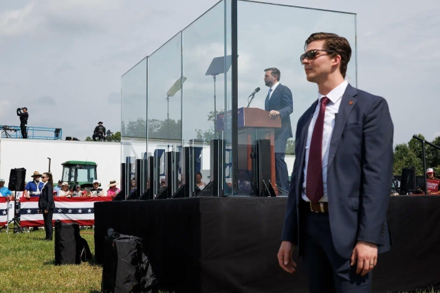 JD Vance on a stage behind bulletproof glass with Secret Service agents flanking him