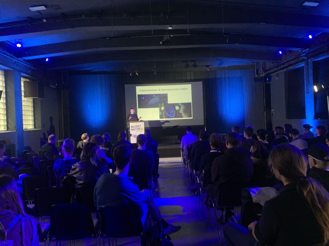 A speaker presents on stage in a dimly lit venue, with an audience seated in rows. The presentation includes slides about databases and biometric data. The setting features blue lighting and a modern aesthetic.