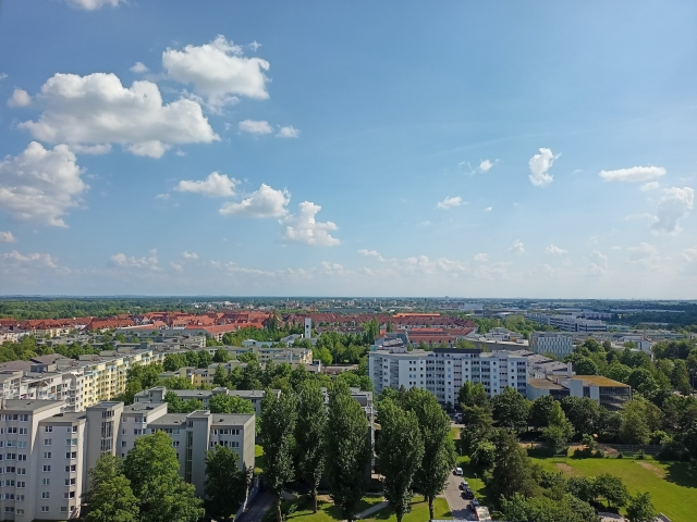 Ausblick aus einem Hochhaus über Grünanlagen und Wohngebäude. Der Himmel ist strahlend blau und leicht bewölkt.