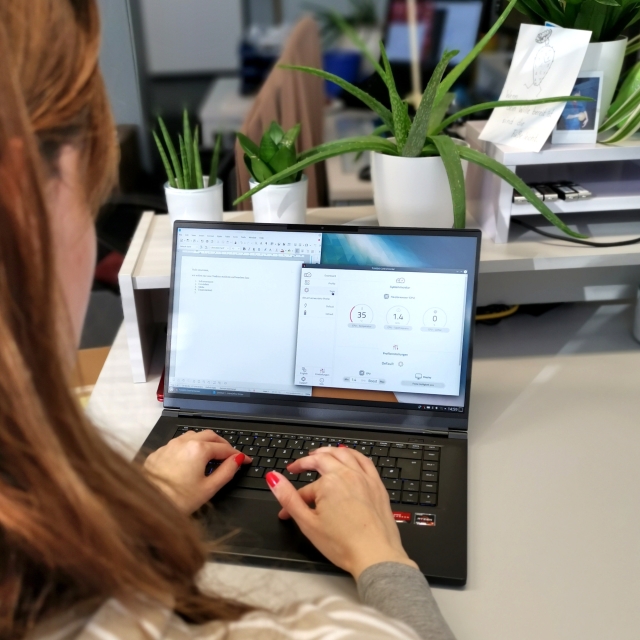 Eine Person mit schulterlangen Haaren sitzt vor einem Notebook, die Hände auf der Tastatur. Im Hintergrund sind Grünpflanzen und Büroeinrichtung zu sehen. Auf dem Bildschirm verschiedene Programmfenster.