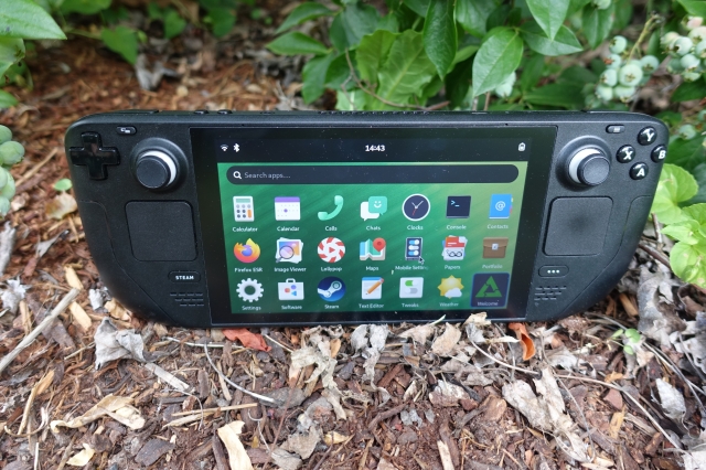 A photo of the Steam Deck handheld gaming console outside sitting on wood chips in front of a blueberry bush. There is a small cluster of blueberries on the upper right, they are unripe and green in color. The Steam Deck's screen is showing the Phosh user interface in landscape mode, with the application launcher menu open. The postmarketOS "welcome" application icon is selected.